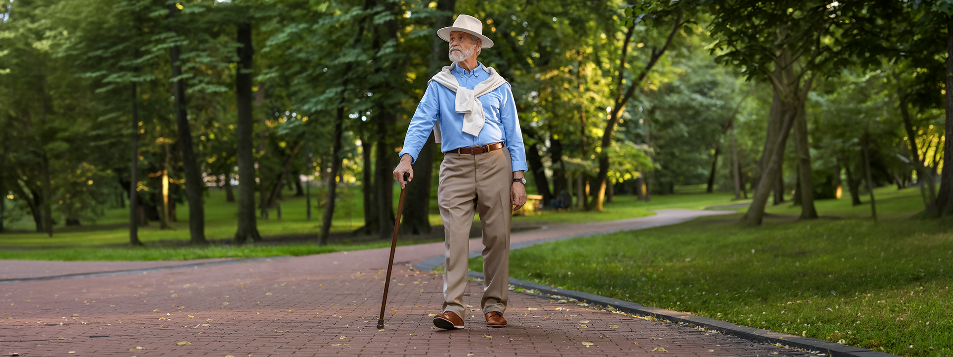 An elderly man walking on a paved path in a park, wearing a light blue button-up shirt, beige trousers, a white bandana, and a beige hat, while holding a walking cane.