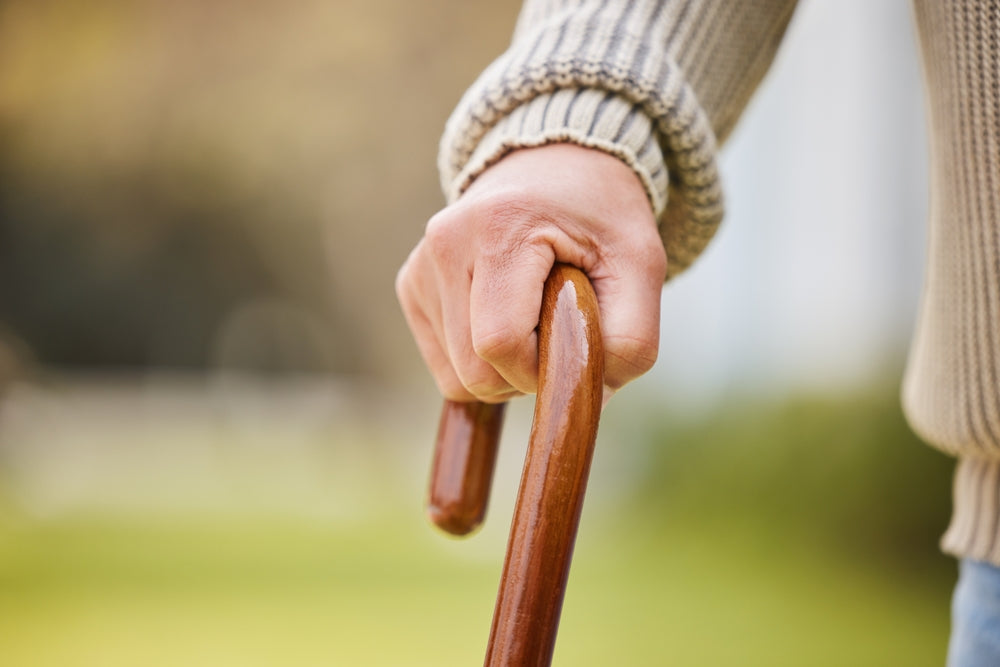 A close-up of a person's hand gripping a wooden cane outdoors, wearing a beige ribbed knit sweater against a blurred green and brown background.