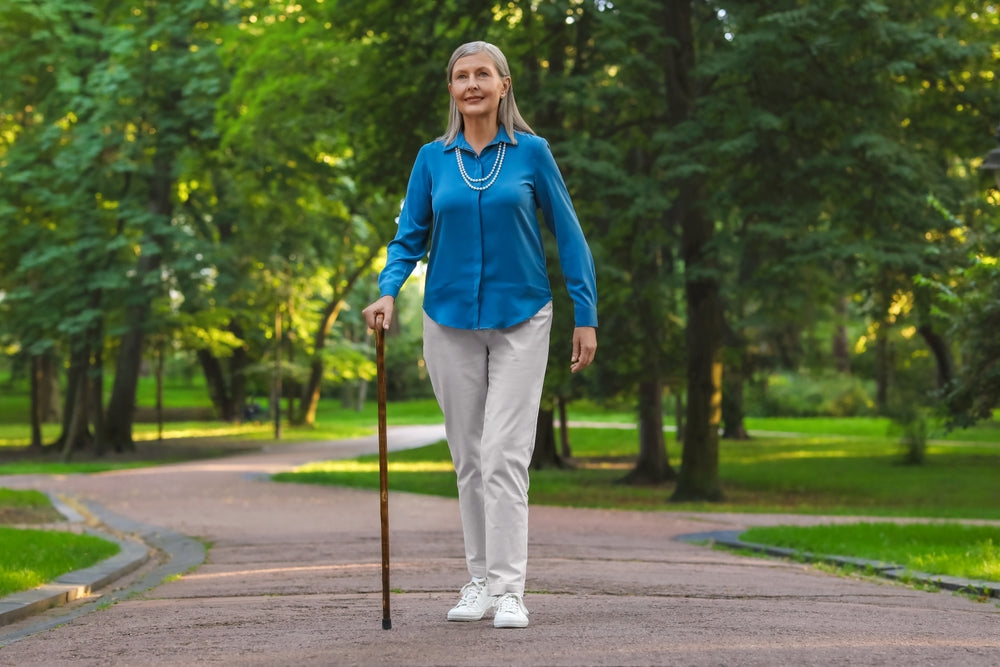 An older woman walking on a paved path in a lush green park, holding a wooden cane. She is wearing a bright blue long-sleeve button-up shirt, light beige trousers, white sneakers, and a layered blue beaded necklace.