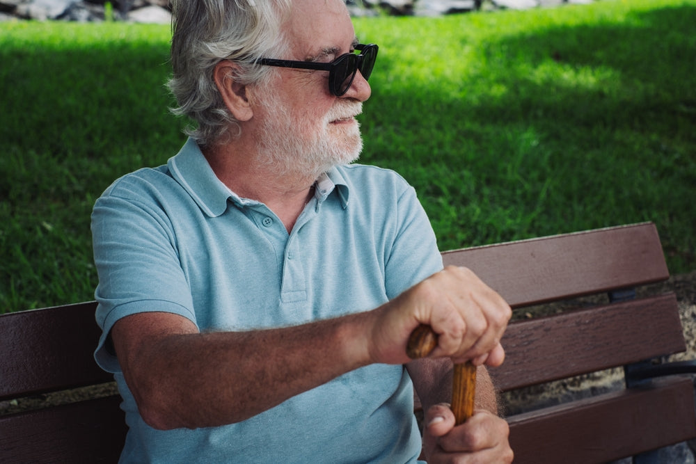 An older man with gray hair and a white beard sits on a wooden park bench, wearing black sunglasses and a light blue polo shirt. He is holding a wooden walking cane in a sunny outdoor setting with green grass in the background.